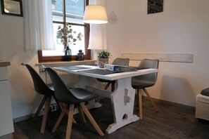 Un coin repas avec une table blanche, deux chaises noires, une lampe et une fenêtre agrémentée d’une plante.