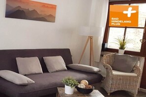 A living room with a dark sofa, a wicker chair, a white lamp, and a painting on the wall. - Holiday apartment with 2 bedrooms (Bad Hindelang)