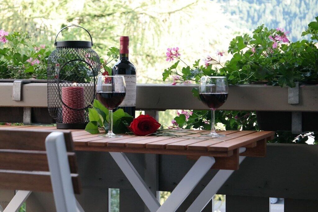 A wooden table with two wine glasses and a bottle, a red rose, and a lantern on a balcony with plants and flowers in the background.