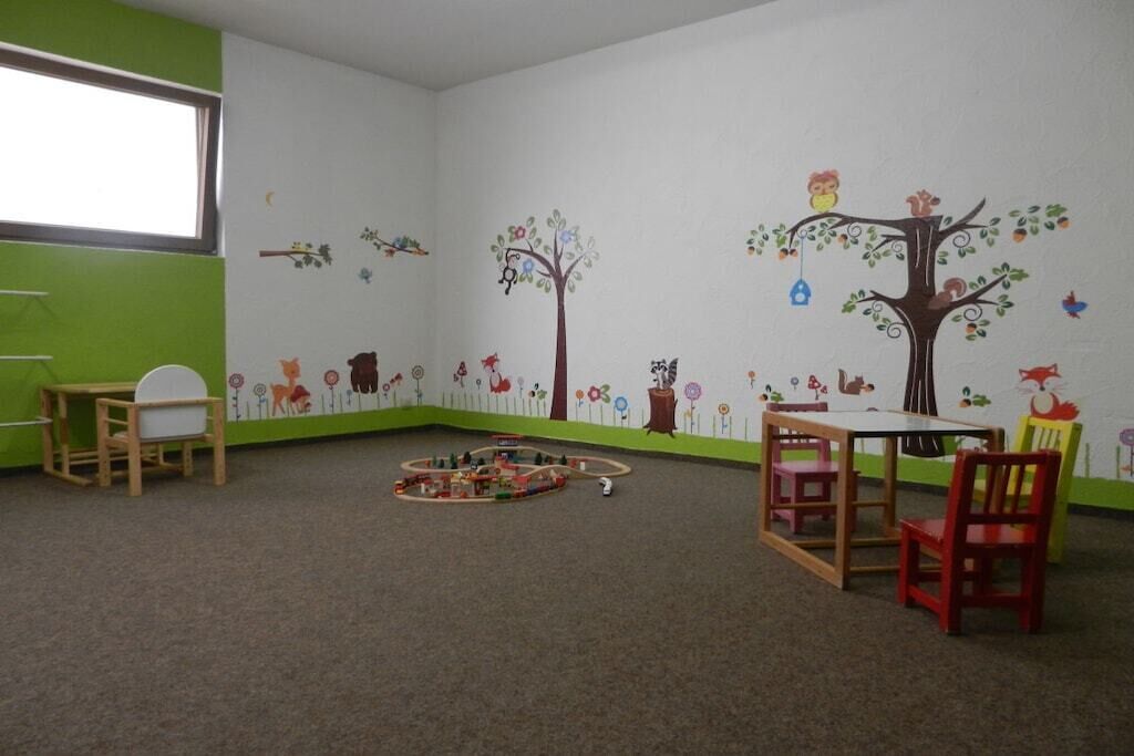 Una stanza con una parete verde, un soffitto bianco, una finestra, un tavolo con sedie e un'area giochi piena di giocattoli.