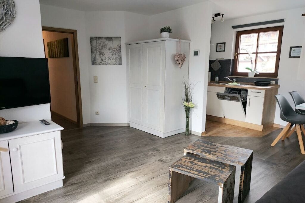 Un espace de vie moderne comprenant un téléviseur à écran plat, un meuble blanc, une table basse en bois, un coin cuisine avec une fenêtre et une armoire blanche.