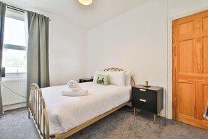Uma cama bem arrumada com um edredom branco, uma mesa de cabeceira com abajur e um guarda-roupa de madeira.