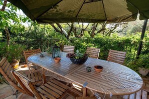 Un área de comedor al aire libre con una mesa redonda, sillas de madera, un paraguas grande y plantas en macetas.