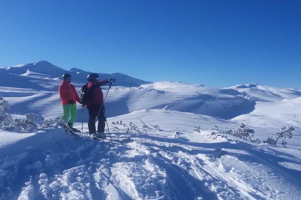 To skiløbere på et snækkedækket bjerg under en klar himmel.