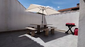 Een terras met een tafel en stoelen, een witte parasol en een rode gasfles.