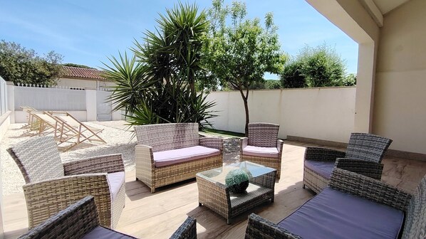 Een terras buiten met rieten meubels, een glazen bijzettafeltje en een grote potplant.