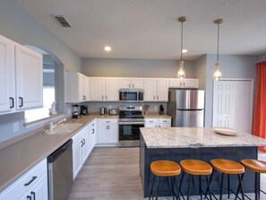 A modern kitchen with white cabinetry, stainless steel appliances, and a granite island with bar stools. - Palms & Pixie Dust Villa Pool (Davenport)