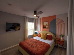 A bedroom with a bed, a ceiling fan, a nightstand, a bedside table, and a wall with a painting. - Palms & Pixie Dust Villa Pool (Davenport)