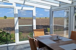 Un invernadero con una mesa y sillas de comedor, puertas de cristal grandes y vistas a un campo arado.
