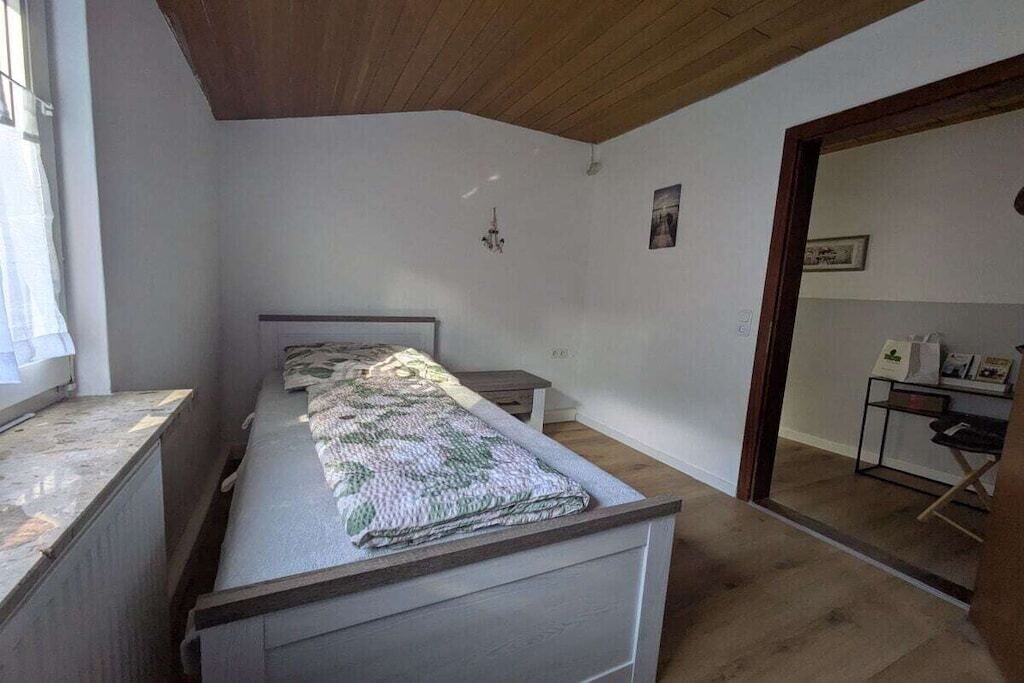 Un dormitorio con una cama, una mesita y una silla. Hay una ventana a la izquierda y una puerta que da a otra habitación.