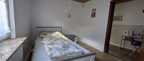 Un dormitorio con una cama, una mesita y una silla. Hay una ventana a la izquierda y una puerta que da a otra habitación.