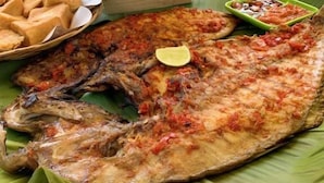 A large grilled fish on a banana leaf with a side of fried items and a small bowl of sauce.
