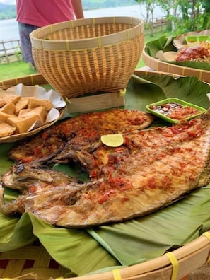 A large grilled fish on a banana leaf with a side of fried items and a small bowl of sauce.