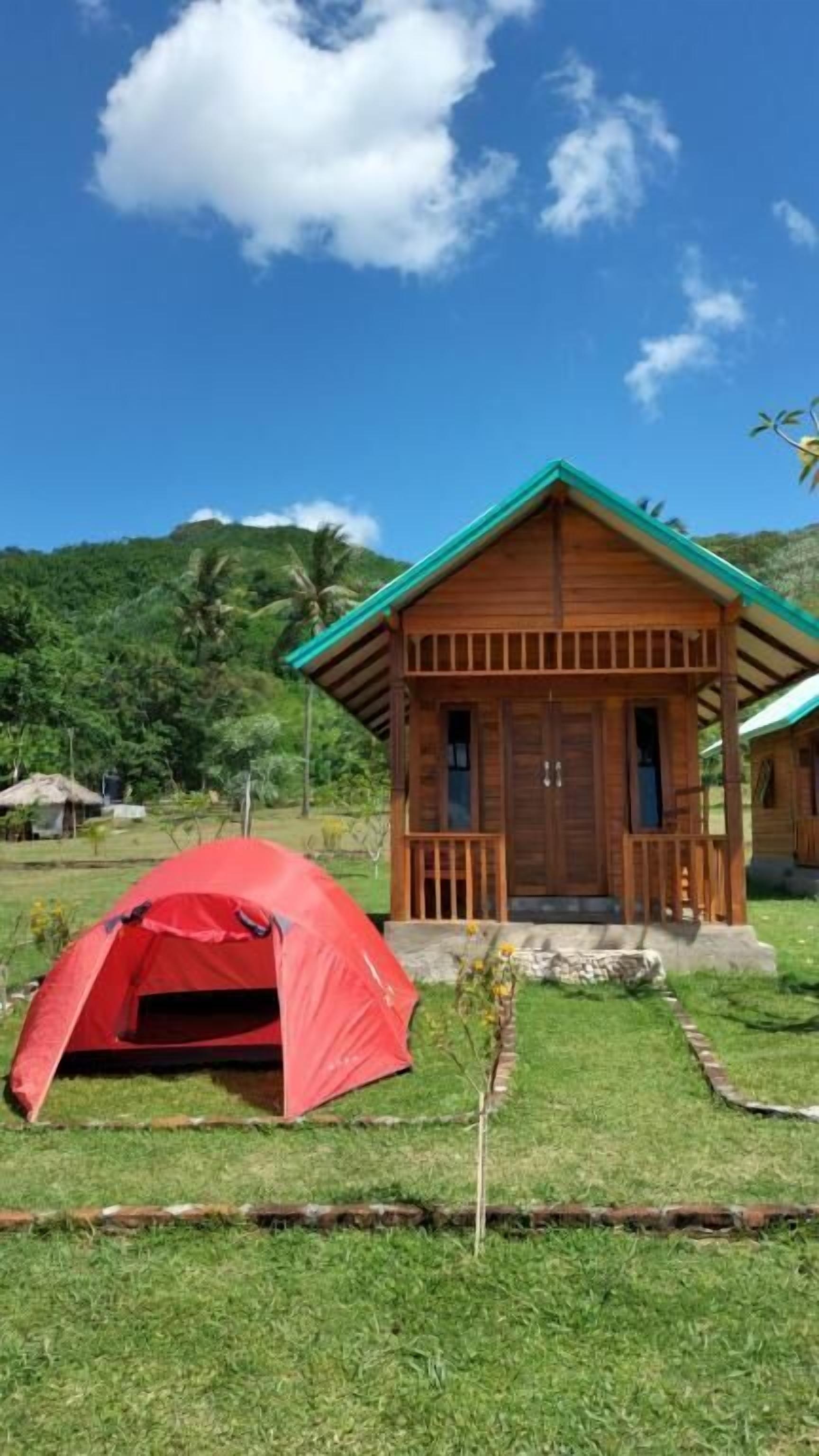 A small wooden cabin with a green roof, a red tent, and a grassy area with a clear sky.