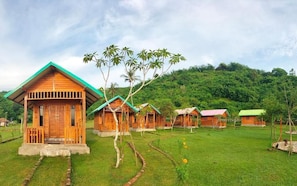 A row of wooden cabins with green roofs, surrounded by lush greenery and a hilly backdrop. - Beach Camp Lombok (Sekotong Barat)