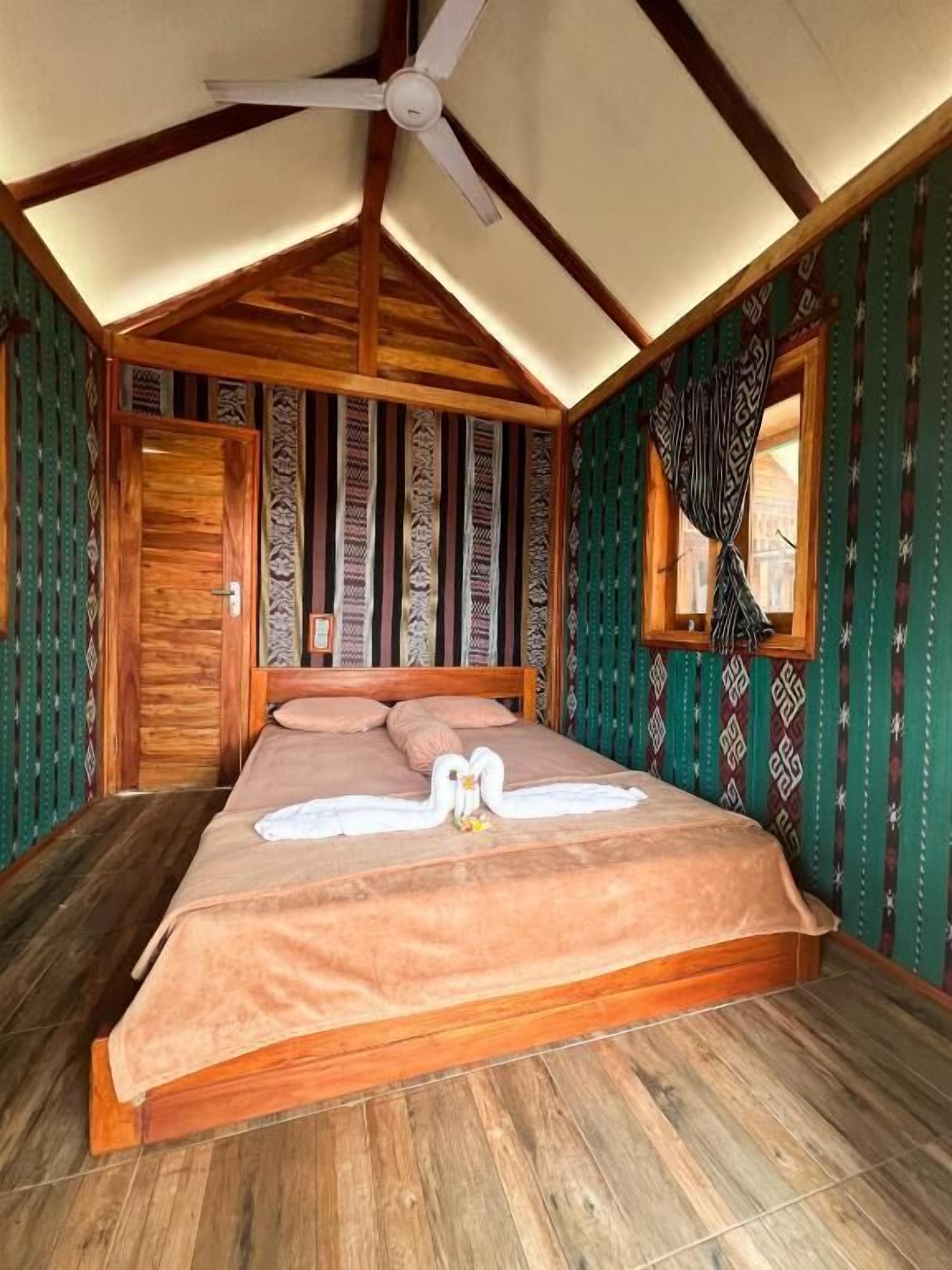 Una habitación con una cama, vigas de madera en el techo, una ventana con cortinas estampadas y una puerta.