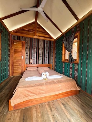 A room with a bed, wooden ceiling beams, a window with patterned curtains, and a door. - Beach Camp Lombok (Sekotong Barat)