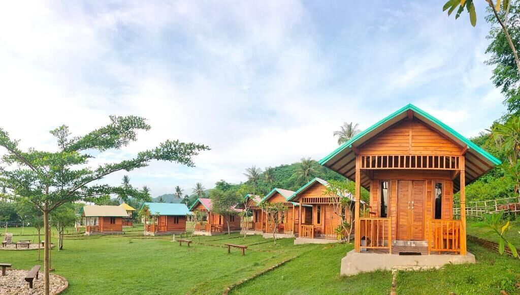 A row of wooden cabins with a green roof, surrounded by a grassy area with trees and a clear sky.
