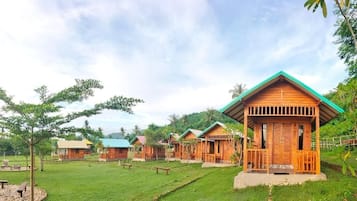 A row of wooden cabins with a green roof, surrounded by a grassy area with trees and a clear sky.
