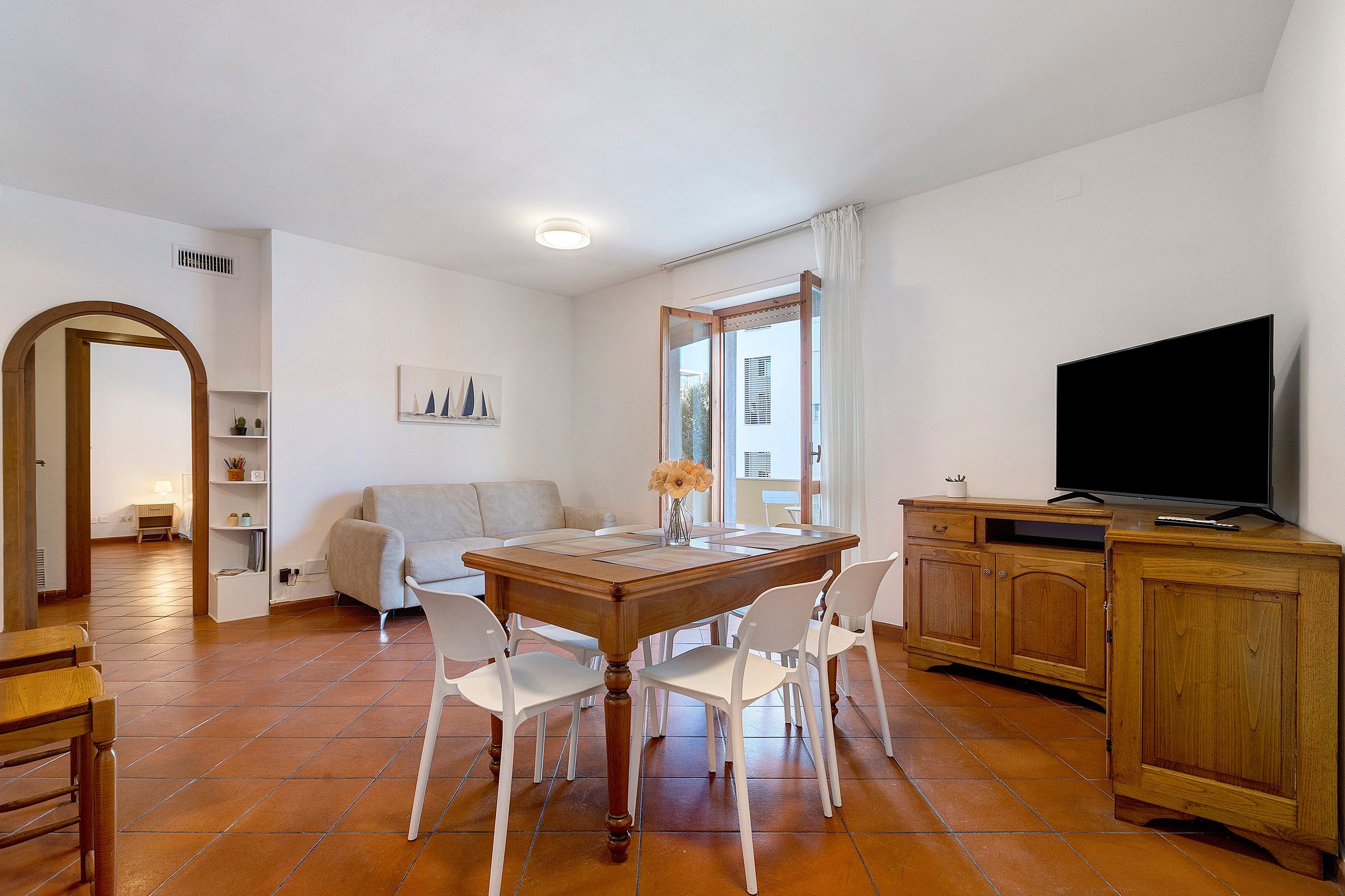 Un comedor con una mesa de madera, sillas blancas, un televisor sobre un mueble de madera y un sofá al fondo.