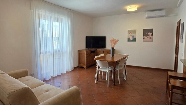 Een woonkamer met een bank, een eettafel met stoelen, een televisie en een keukenhoek met een airconditioner.