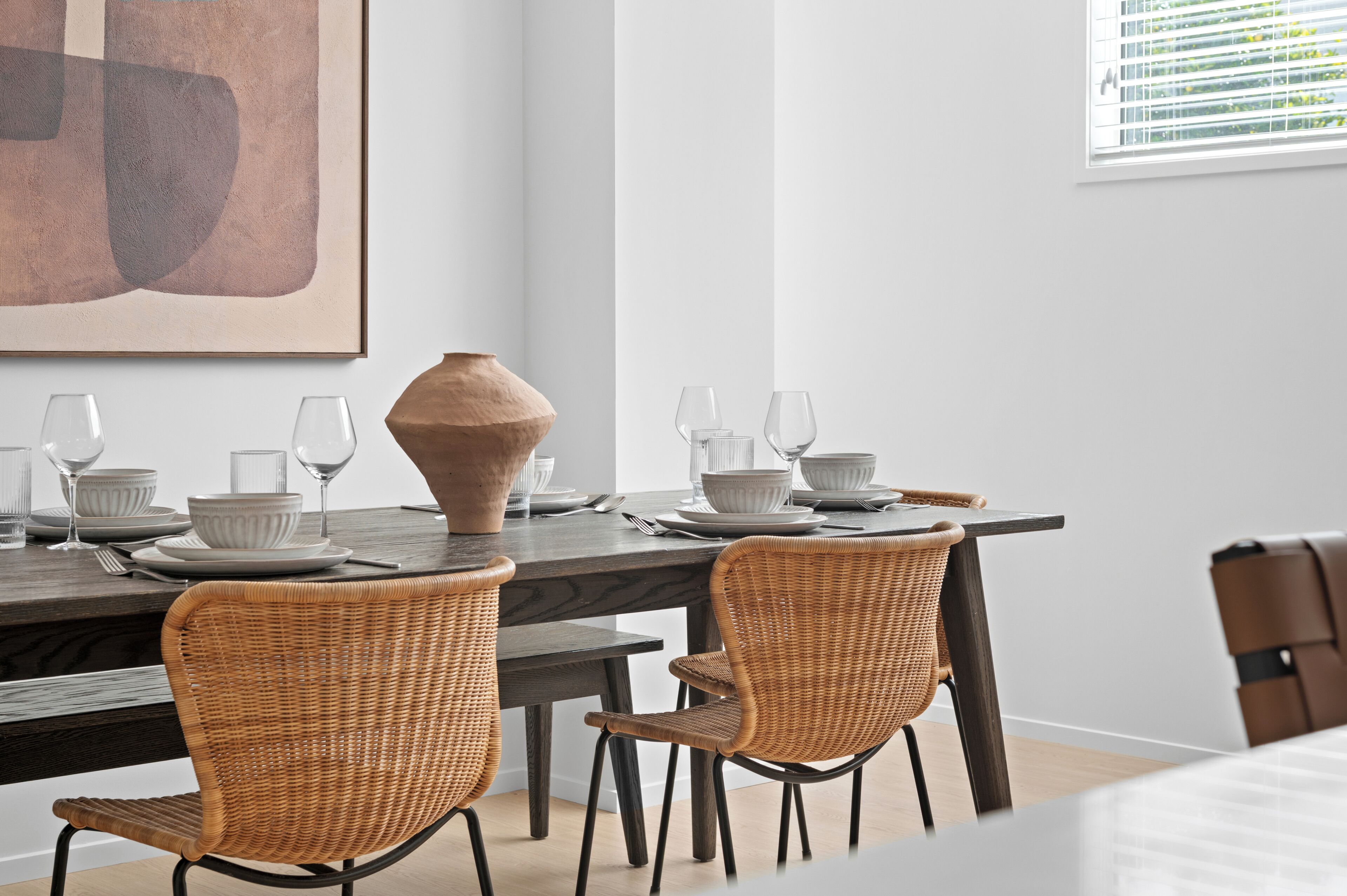 A dining area with a dark wooden table, white plates, and clear glassware. There are wicker chairs and a large vase on the table.