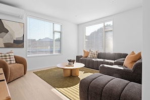 Een moderne woonkamer met een hoekbank, een ronde salontafel en een groot raam met zonwering.