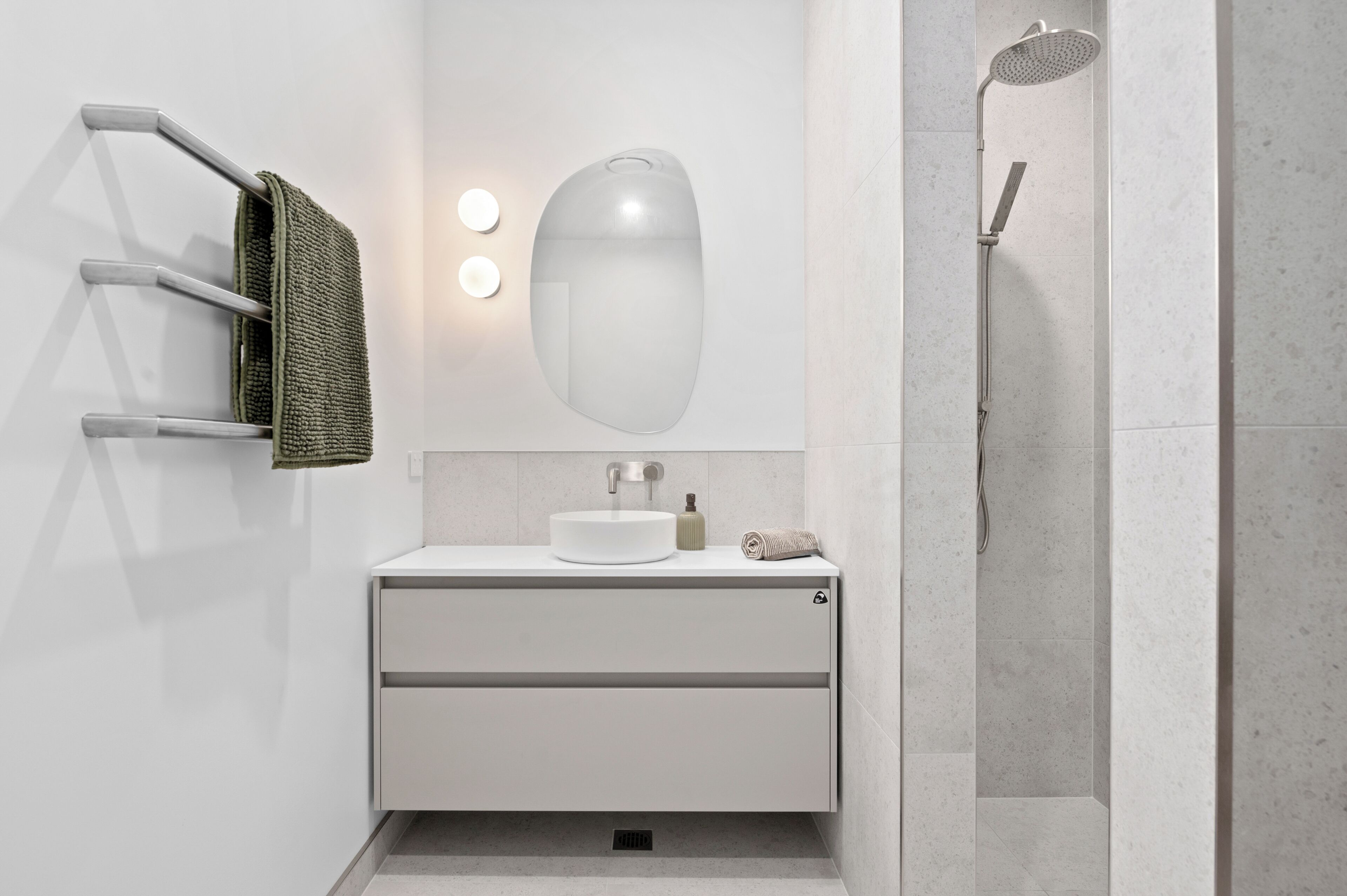 A modern bathroom with a white vanity, a round mirror, a towel rack, and a shower area.