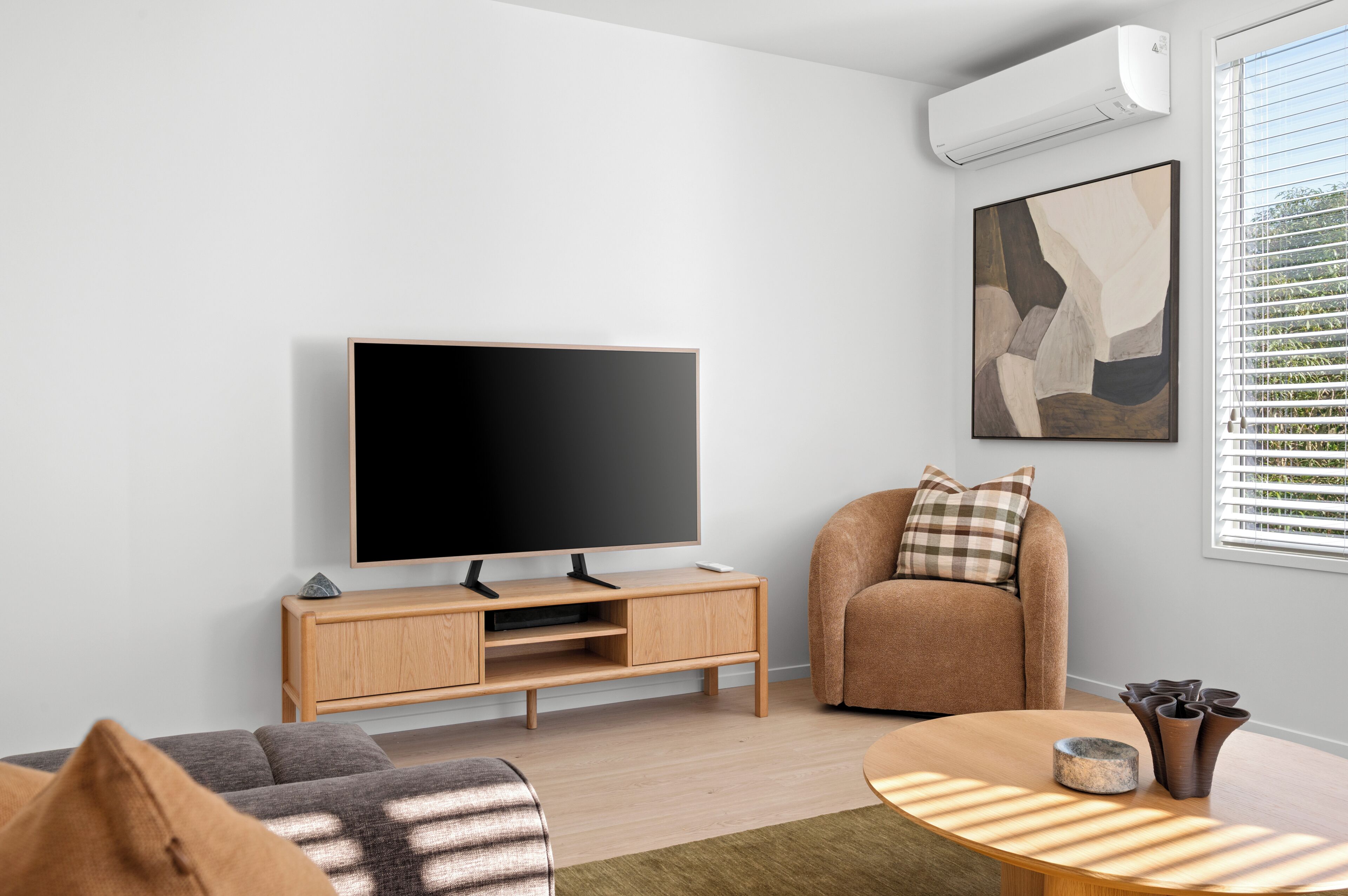 A modern living room with a flat-screen TV, a wooden entertainment unit, a round coffee table, a sofa, and a window with blinds.