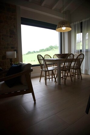 Ein Essbereich mit Tisch und Stühlen, einer Hängelampe und Blick auf eine grüne Landschaft durch große Fenster.