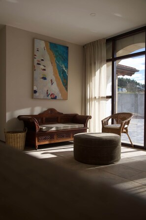 Ein Zimmer mit einem großen Strandgemälde, einem Holzsofa, einem Korbstuhl und einem großen Fenster mit Vorhängen.