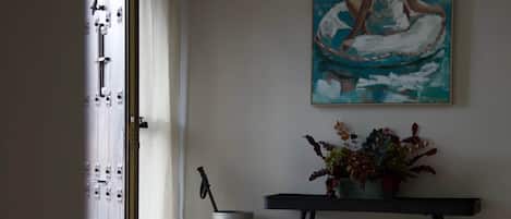 A room with a large painting of a woman in a white dress, a small table with a vase of flowers, and a metal container on the floor.