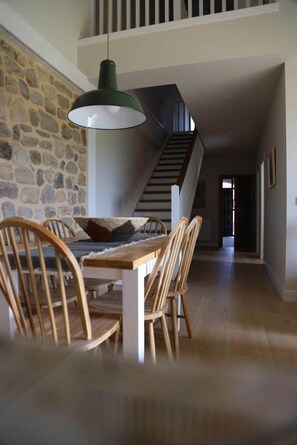 A dining area with a stone wall, wooden chairs, a table, and a hanging light fixture. - Sierra Pando. Valdhaya Homes (Cantabria)