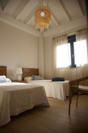 Ein Schlafzimmer mit zwei Betten, einem Holzkopfende, einem Korbstuhl, einem Fenster mit transparenten Vorhängen und einer gewebebespannten Deckenleuchte.