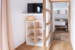 Une pièce avec une télévision posée sur une étagère, un chapeau également posé sur une étagère, et une salle de bain avec un miroir et un lavabo.