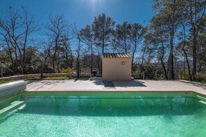 Ein Schwimmbecken mit einer klaren blauen Wasseroberfläche, umgeben von einer Betonplattform und einem kleinen Gebäude mit einem Ziegeldach.