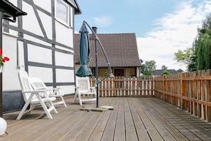 Une terrasse en bois avec des chaises blanches et une table, une clôture en bois et un bâtiment doté d’un toit en tuiles.