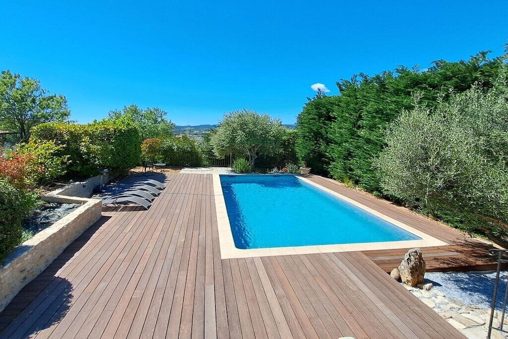 A rectangular swimming pool with a wooden deck, surrounded by various trees and shrubs.