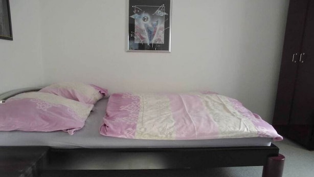 A single bed with a pink and white bedspread, a framed abstract artwork on the wall, and a dark-colored wardrobe.