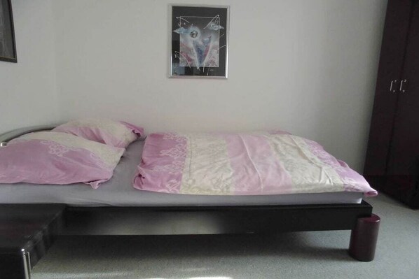 A single bed with a pink and white bedspread, a framed abstract artwork on the wall, and a dark-colored wardrobe.