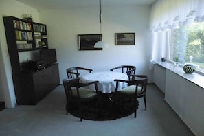 A room with a dining table surrounded by chairs, a bookshelf, a television, and a window with curtains.