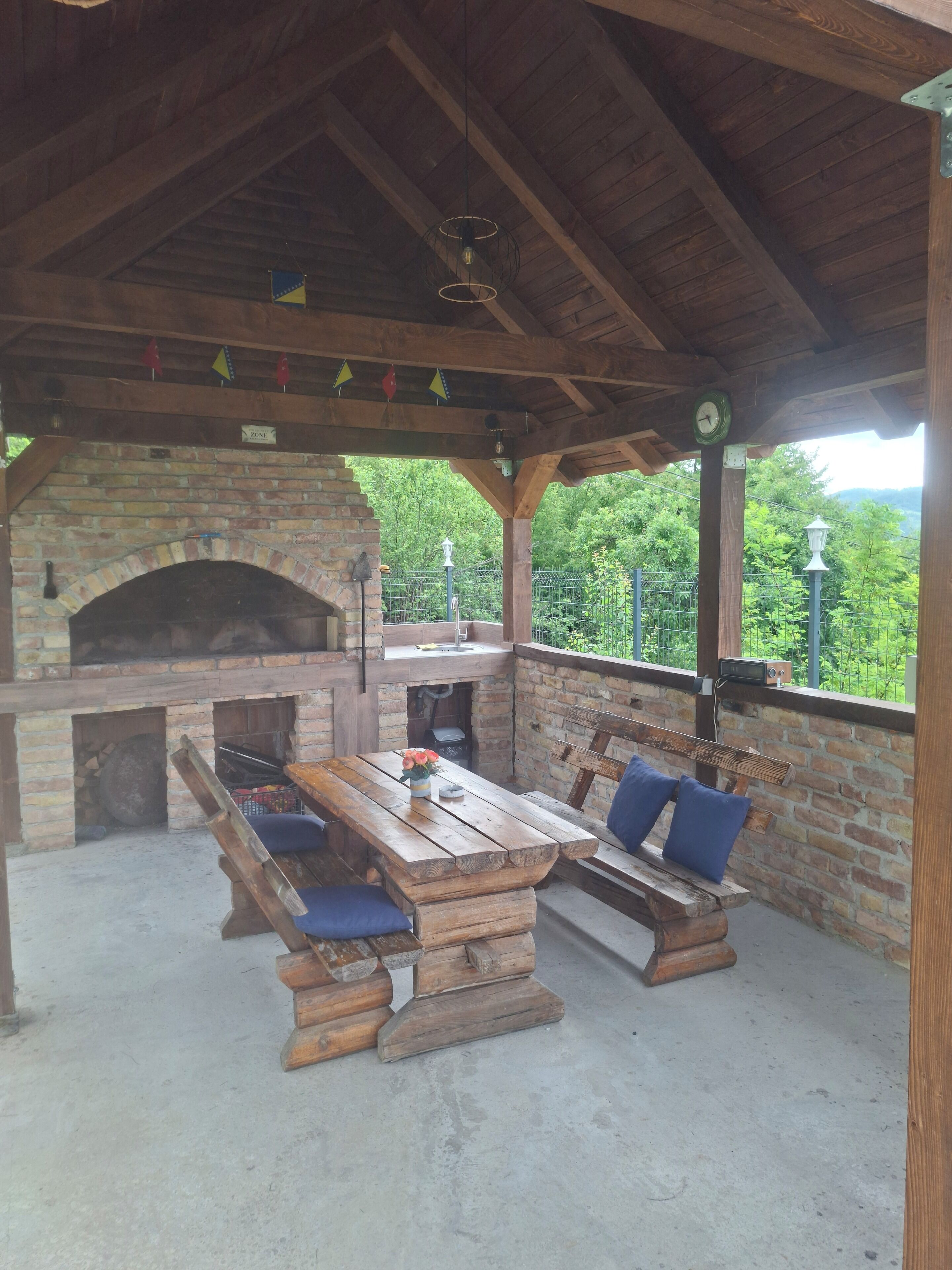 Une terrasse extérieure avec une cheminée en briques, des bancs en bois et une table agrémentée d’un vase de fleurs.