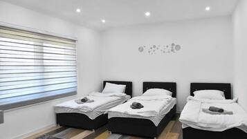 A room with three beds, a window with blinds, a wall-mounted light fixture, and a patterned rug.