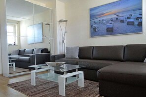 A modern living room with a brown sofa, a glass coffee table, and a painting of a beach scene. - Villa Parkblick Apt. 17 (Kühlungsborn)