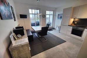 A modern kitchen with a built-in oven, a dining area with a table and chairs, and a large window providing natural light. - Villa Parkblick Apt. 17 (Kühlungsborn)