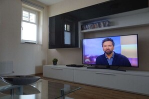 A modern living room with a flat-screen TV mounted on a white entertainment unit, a glass coffee table, and a window with blinds. - Villa Parkblick Apt. 17 (Kühlungsborn)