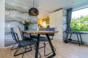 Un comedor con mesa de madera, cuatro sillas, una lámpara colgante y vistas a una cocina y vegetación exterior.
