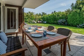 An outdoor patio area with a wooden table set for dining, surrounded by wicker chairs, and a cushioned sofa. - "t Ongerepte 56 - De Zandbank (Renesse)