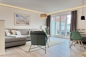Un salon moderne comprenant un canapé, un fauteuil vert, une table à manger avec des chaises et une grande fenêtre donnant sur des immeubles.