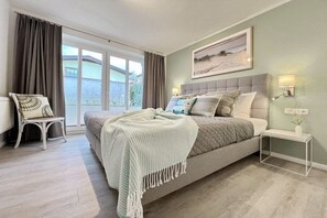 Un dormitorio con una cama grande, una mesita de noche, una silla y un ventanal con cortinas.